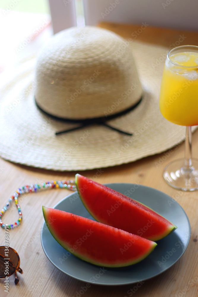 Plate with watermelon slices, glass of orange juice or cocktail, straw hat, colorful beaded necklace and sunglasses on the wooden table. Summer essentials concept. Selective focus.