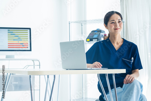 A female nurse in blue scrubs works on a laptop at a medical desk, with a digital bar chart displayed on the screen behind her. The setting is a bright, modern hospital or clinic office