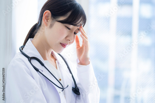 A female doctor in a white coat touches both her temples with her hands, eyes closed, appearing to suffer from a headache or stress. A stethoscope is visible around her neck.

