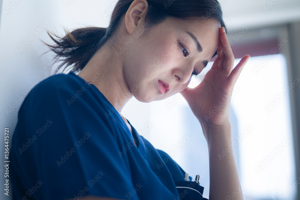 A close-up of a nurse in blue scrubs holding her forehead, eyes closed, showing signs of mental fatigue or stress in a clinical environment.