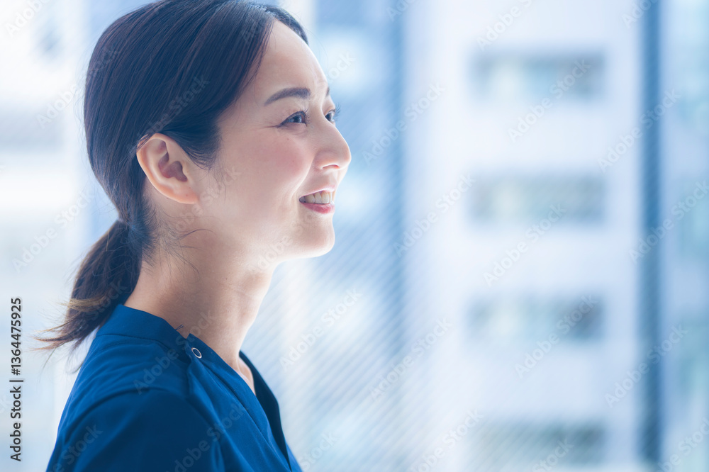 A smiling female nurse in blue scrubs looks out a bright hospital window. The expression contrasts previous images, representing hope, resilience, and optimism.