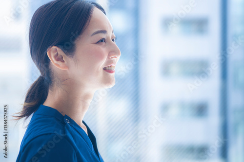 A smiling female nurse in blue scrubs looks out a bright hospital window. The expression contrasts previous images, representing hope, resilience, and optimism.