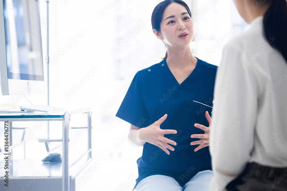 A female doctor wearing blue scrubs talks to a female patient while gesturing with both hands during a medical consultation in a modern clinic.