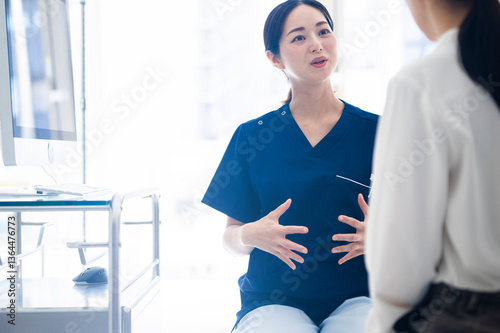 Obraz na płótnie A female doctor wearing blue scrubs talks to a female patient while gesturing with both hands during a medical consultation in a modern clinic. w salonie
