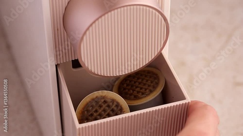 A woman's hand shows the compartment for used capsules from a capsule coffee machine in pale beige, champagne or rose gold. Recyclable plastic coffee capsules with a paper lid