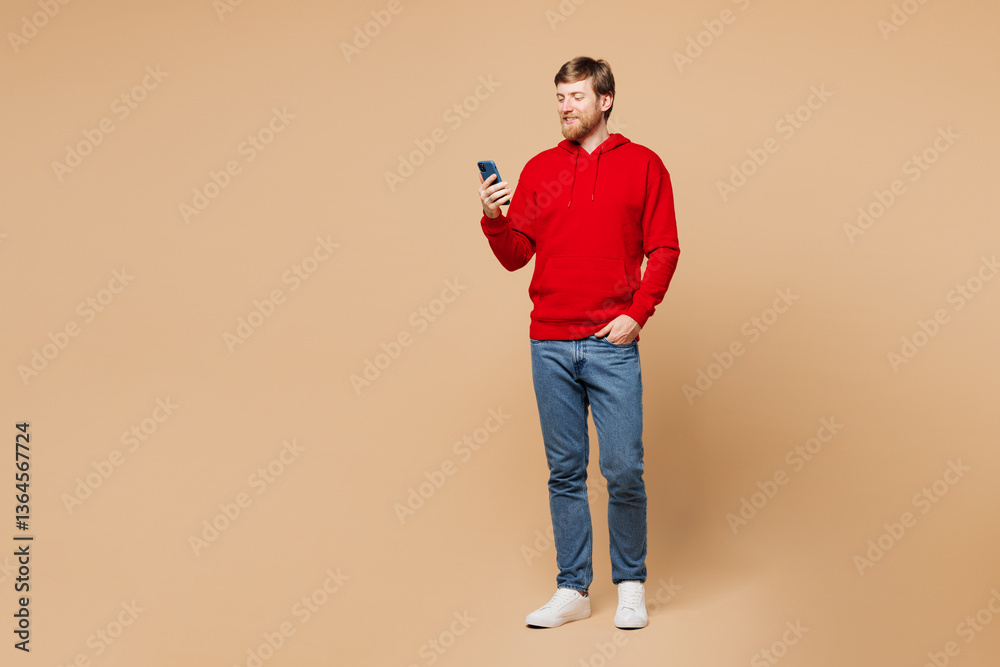 Fototapeta premium Full body young smiling happy man wear red hoodie casual clothes hold in hand use mobile cell phone typing message chat online isolated on pastel plain light beige background studio Lifestyle concept