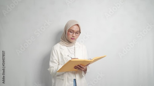A asian woman in a hijab and glasses, holding a document with a pen.