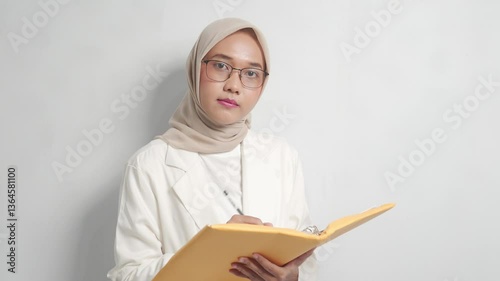 A asian woman wearing a hijab, blazer, and glasses, writing in a yellow folder.