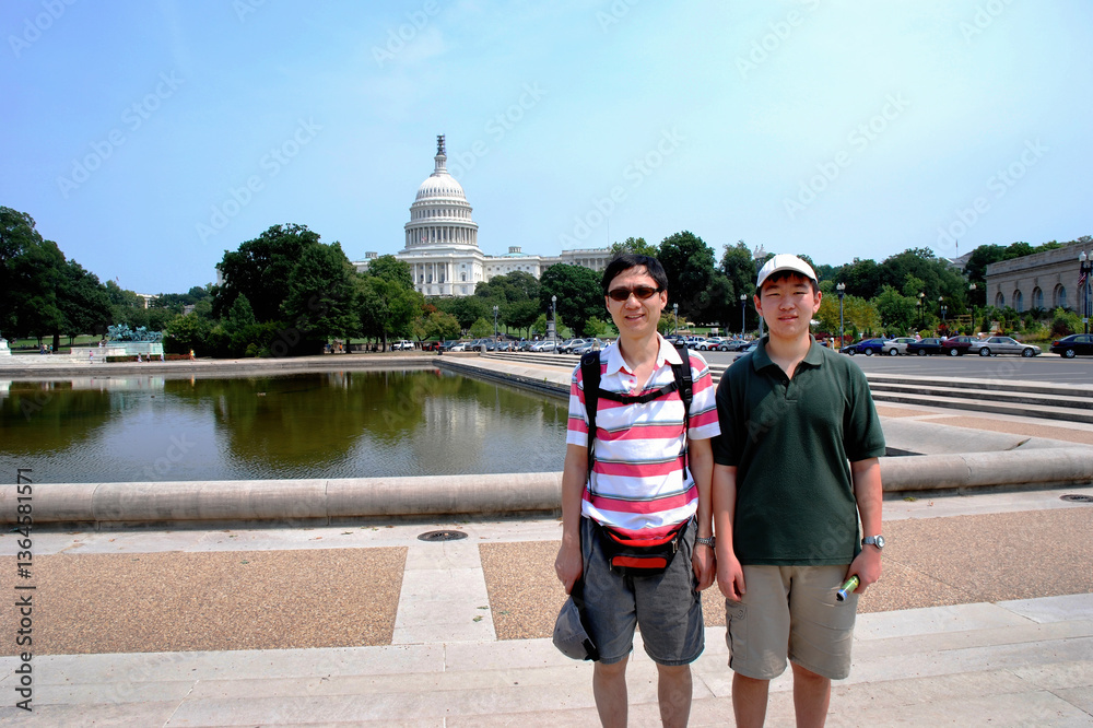 Tourists posing for photos with Capitol Hill in the distance. Unrecognizable cars and crowds in the background. Washington D.C.