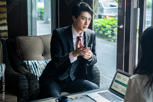 A businessman in a suit uses expressive hand gestures while explaining a concept to his female colleague in a business meeting at a café. The laptop screen is visible.