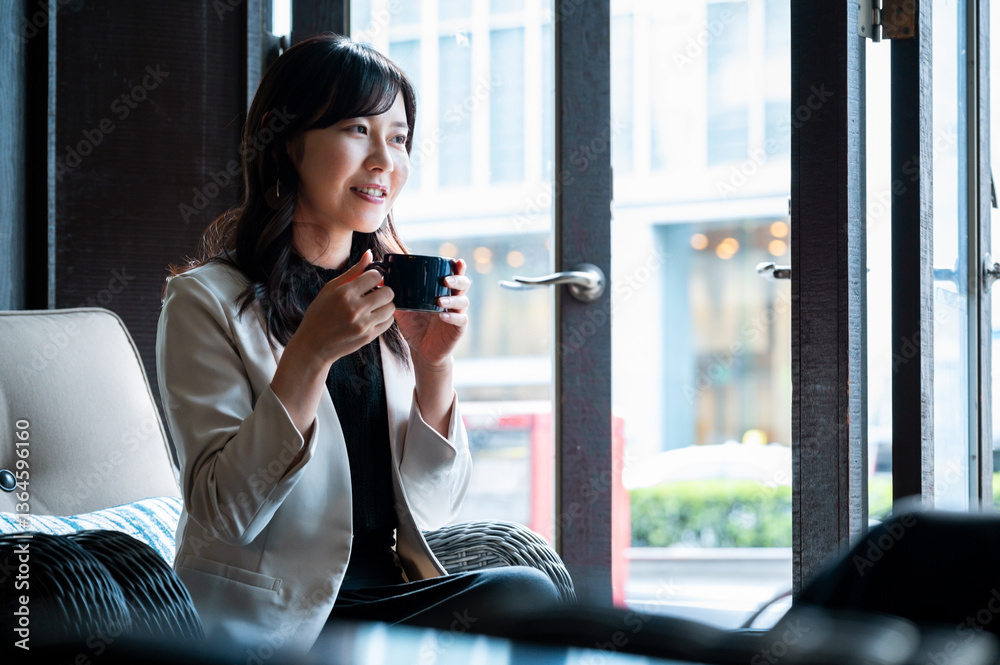 cheerful woman in a cream blazer enjoys a cup of coffee while seated by a window in a stylish café. Natural daylight highlights her relaxed mood as she looks toward someone outside the frame.