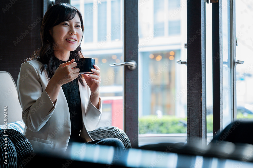 cheerful woman in a cream blazer enjoys a cup of coffee while seated by a window in a stylish café. Natural daylight highlights her relaxed mood as she looks toward someone outside the frame.