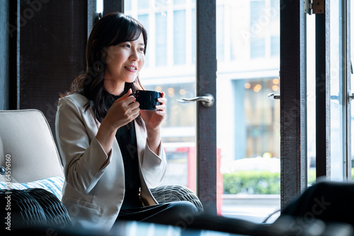 Obraz na płótnie cheerful woman in a cream blazer enjoys a cup of coffee while seated by a window in a stylish café. Natural daylight highlights her relaxed mood as she looks toward someone outside the frame.
w salonie