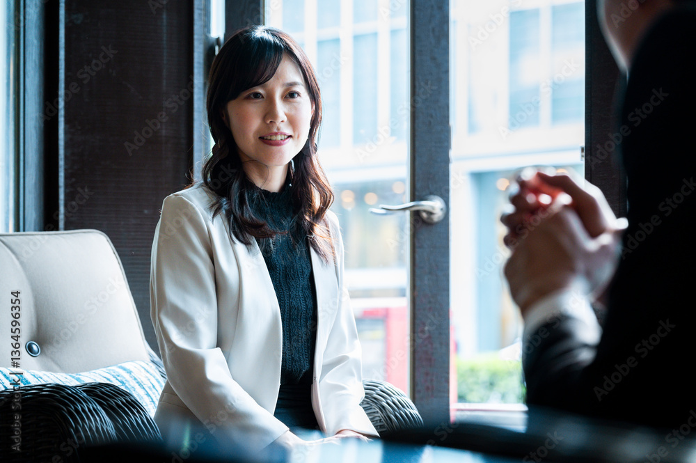 A woman dressed in a cream-colored blazer attentively listens to someone across from her during a relaxed conversation in a modern café. The soft daylight from the window illuminates her face.