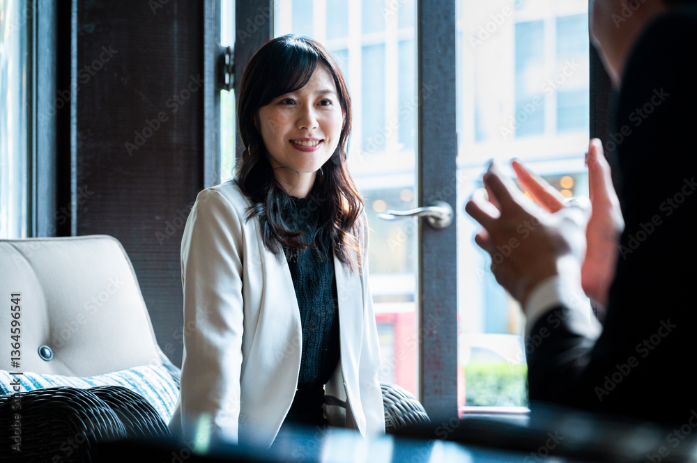 A woman dressed in a cream-colored blazer attentively listens to someone across from her during a relaxed conversation in a modern café. The soft daylight from the window illuminates her face.