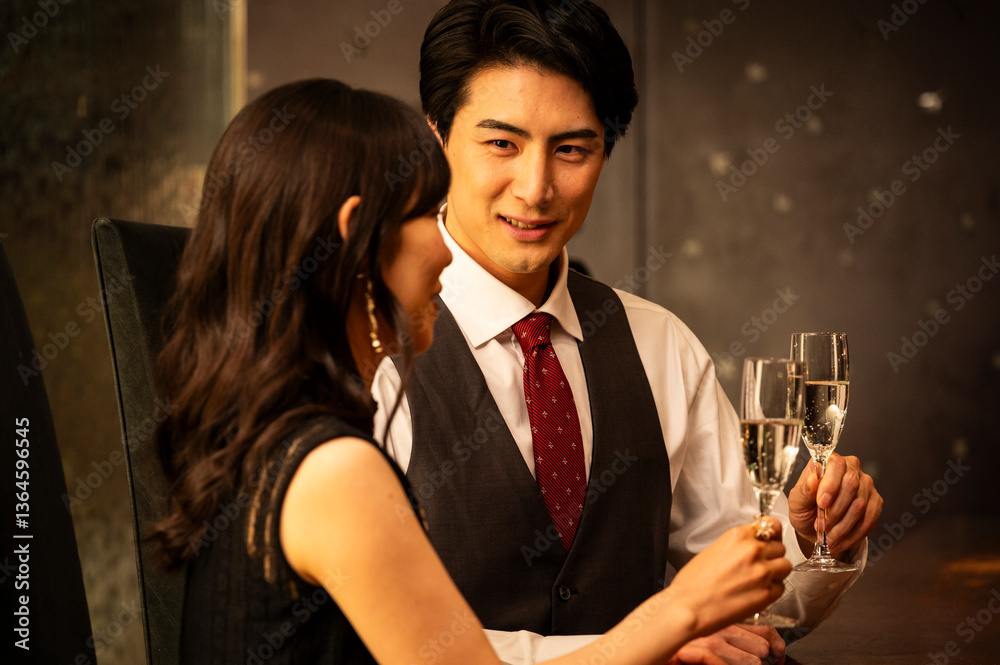 A well-dressed man and woman toast with champagne glasses while sitting at a modern upscale bar. The man wears a white shirt with a dark vest and red tie, and the woman is dressed in a black sleeveles