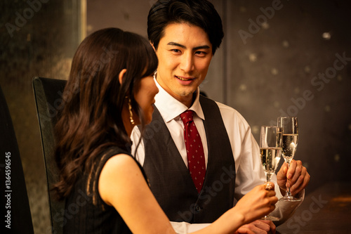 Obraz na płótnie A well-dressed man and woman toast with champagne glasses while sitting at a modern upscale bar. The man wears a white shirt with a dark vest and red tie, and the woman is dressed in a black sleeveles w salonie