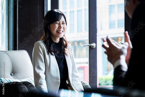 A woman dressed in a cream-colored blazer attentively listens to someone across from her during a relaxed conversation in a modern café. The soft daylight from the window illuminates her face.