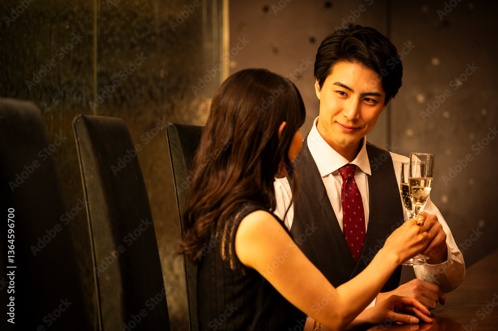 A well-dressed man and woman toast with champagne glasses while sitting at a modern upscale bar. The man wears a white shirt with a dark vest and red tie, and the woman is dressed in a black sleeveles