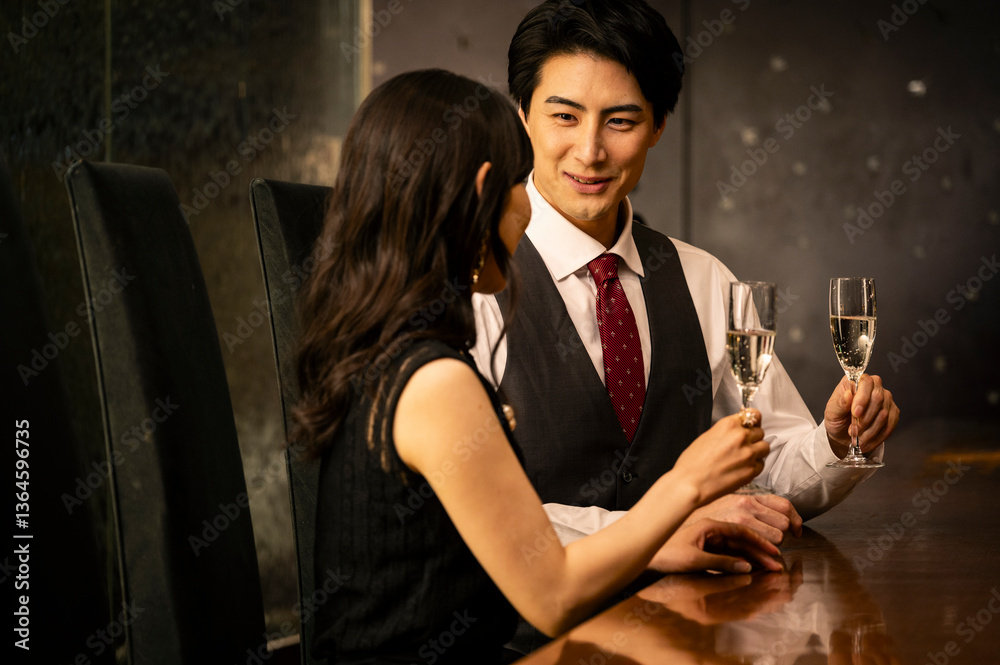 A well-dressed man and woman toast with champagne glasses while sitting at a modern upscale bar. The man wears a white shirt with a dark vest and red tie, and the woman is dressed in a black sleeveles