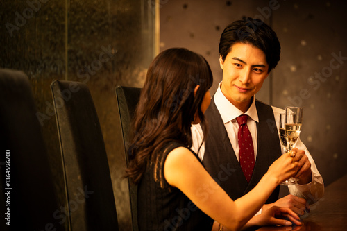 Obraz na płótnie A well-dressed man and woman toast with champagne glasses while sitting at a modern upscale bar. The man wears a white shirt with a dark vest and red tie, and the woman is dressed in a black sleeveles w salonie