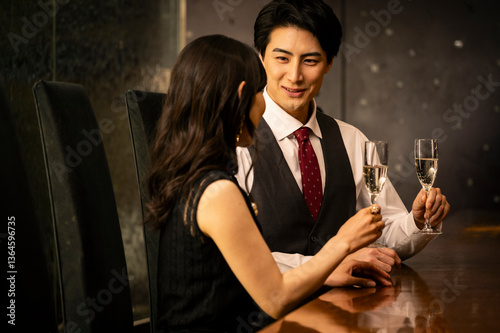 Obraz na płótnie A well-dressed man and woman toast with champagne glasses while sitting at a modern upscale bar. The man wears a white shirt with a dark vest and red tie, and the woman is dressed in a black sleeveles w salonie