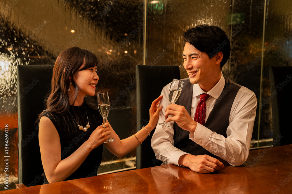 A man and woman dressed formally are smiling at each other while toasting with champagne glasses. They are seated across from each other at a wooden bar with warm ambient lighting.