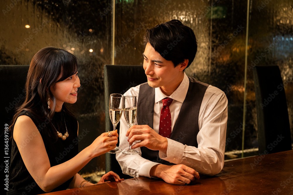 A well-dressed man and woman toast with champagne glasses while sitting at a modern upscale bar. The man wears a white shirt with a dark vest and red tie, and the woman is dressed in a black sleeveles
