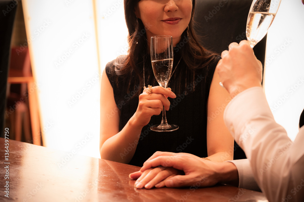 A man in a white shirt and vest and a woman in a black dress toast with champagne glasses at a high-end bar. The couple is smiling and enjoying a relaxed evening in a dimly lit, elegant setting.