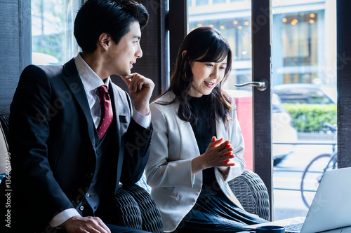 Obraz na płótnie A businessman and a businesswoman in formal attire have a serious conversation while seated by the window at a modern café. Natural light highlights their focused expressions as they engage in a profe w salonie