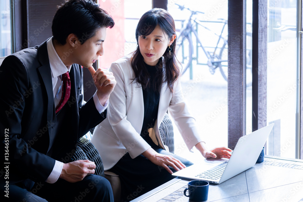 A businessman and a businesswoman in formal attire have a serious conversation while seated by the window at a modern café. Natural light highlights their focused expressions as they engage in a profe