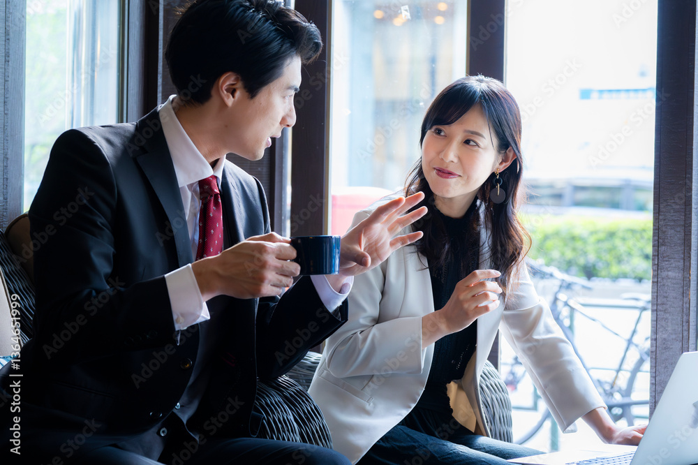 A businessman in a dark suit and red tie gestures while speaking with a female colleague in a light blazer during a strategic meeting in a cozy café with a brick wall background.