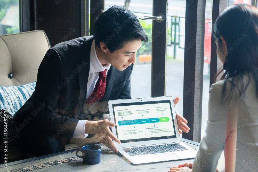 A businessman in formal attire is pointing to a laptop screen and explaining data to a female colleague across the table. They are seated by a bright window in a stylish café setting with a coffee mug