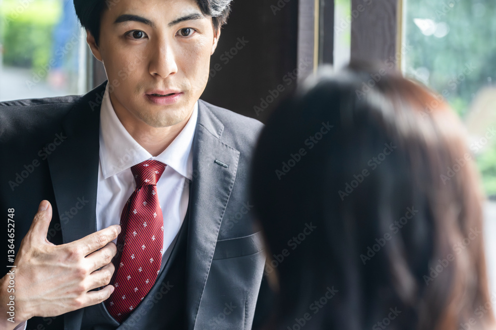 A businessman in formal attire is pointing to a laptop screen and explaining data to a female colleague across the table. They are seated by a bright window in a stylish café setting with a coffee mug