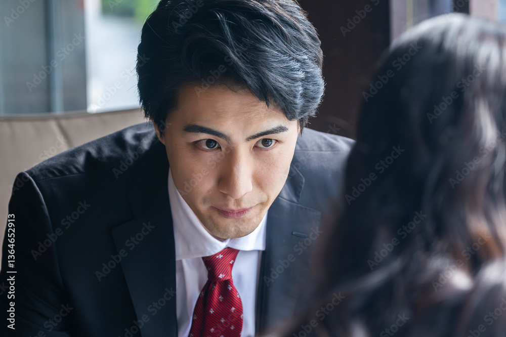 A businessman in formal attire is pointing to a laptop screen and explaining data to a female colleague across the table. They are seated by a bright window in a stylish café setting with a coffee mug