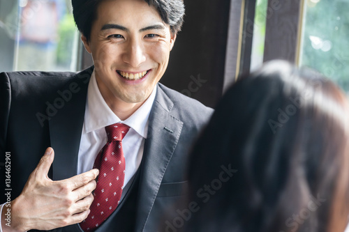 A businessman in formal attire is pointing to a laptop screen and explaining data to a female colleague across the table. They are seated by a bright window in a stylish café setting with a coffee mug
