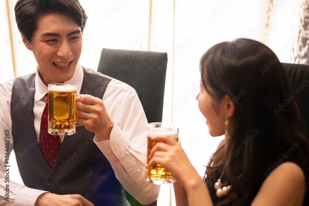 A smiling man and woman enjoy a moment of celebration with beer at a sleek bar. The man wears a white shirt with a red tie and vest, and the woman smiles warmly as they toast.