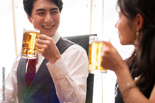 Obraz na płótnie A smiling man and woman enjoy a moment of celebration with beer at a sleek bar. The man wears a white shirt with a red tie and vest, and the woman smiles warmly as they toast. w salonie