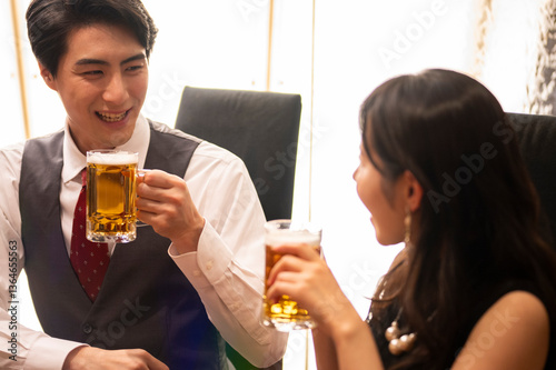 Obraz na płótnie A smiling man and woman enjoy a moment of celebration with beer at a sleek bar. The man wears a white shirt with a red tie and vest, and the woman smiles warmly as they toast. w salonie