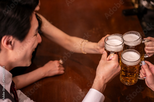 A warm-toned image of 4 people clinking beer mugs at a modern bar. The close-up focuses on their hands and glasses filled with golden beer, capturing a festive group moment.