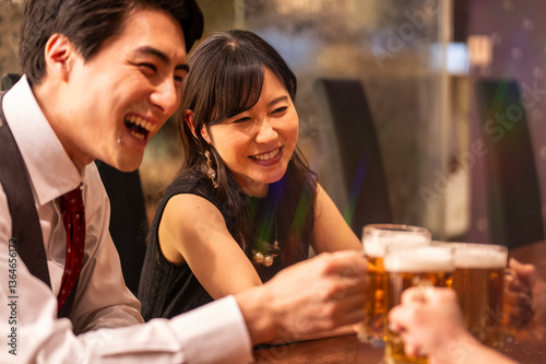 Obraz na płótnie A joyful group of 4 friends toasts with beer mugs while sitting at a polished wooden bar. The ambient lighting and formal clothing suggest a special celebration or social gathering. w salonie