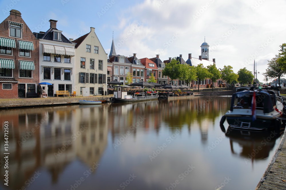 Naklejka premium Reflections of traditional houses, barges and boats on the little canal (klein Diep) in Dokkum, Friesland, Netherlands, with the Oud Stadhuis (Old Town Hall, built in 1610) in the background