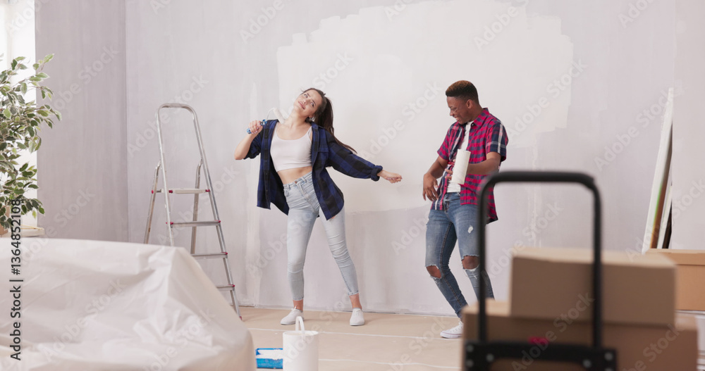 Fototapeta premium Smiling, cheerful friends have fun renovating their apartment, playfully painting walls with rollers. The room is filled with laughter, with furniture securely covered to protect against dirt.
