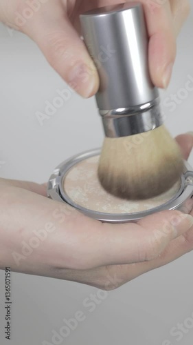 Vertical footage, application of compact powder.The woman is tapping the retractable face brush onto the compact. She picks up the powder with the brush, preparing to apply it to her face.