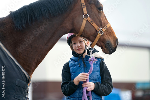 Teenager holds purple lead rope and gently pets horse in riding helmet and blue vest standing. Brown horse and young girl with blanket near wooden stable. Equestrian sport, activity, horse racing.
