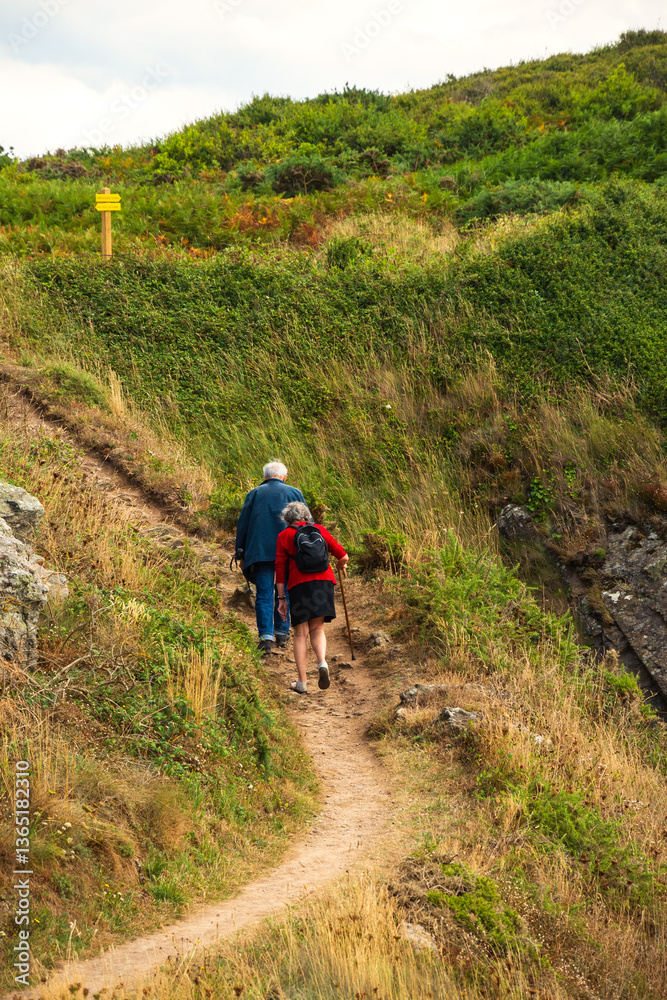 Obraz premium Senior couple (back view) hiking in in summer at picturesque hills in Emerald Coast, Brittany, France. Healthy, active lifestyle, elderly wellbeing concepts. French countryside tourism background.