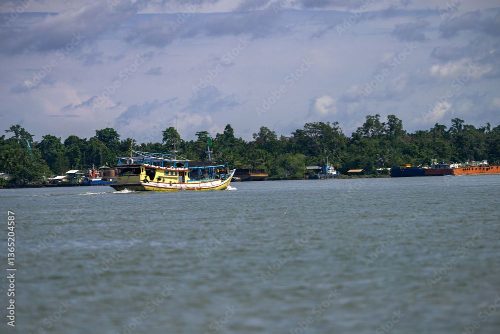 Fototapeta premium A colorful fishing boat sails on a calm river with lush green trees and buildings in the background. Other boats are docked along the shore under a partly cloudy sky