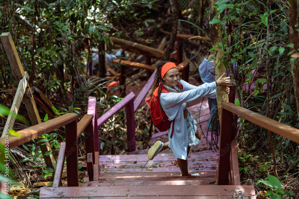 Fototapeta premium A woman with a red headscarf and casual outfit climbs a red wooden staircase in a lush tropical forest, holding the railings. Sunlight filters through the dense green foliage, creating a vibrant atmos