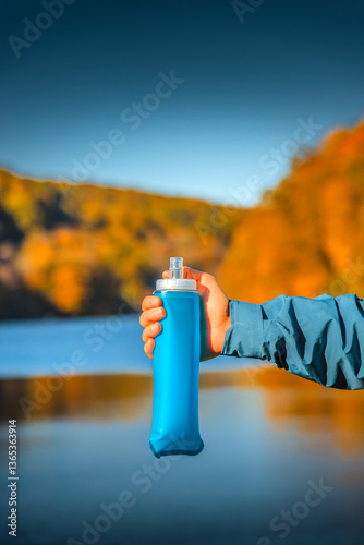 A hiker’s hand holds a blue soft water bottle against a scenic autumn landscape with a lake and colorful foliage.