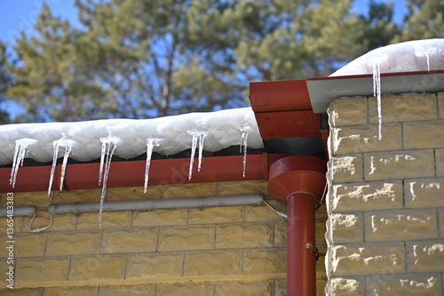 Snow and icicles on the roof of the house against the background of blue sky and pine forest in blur. Sunny spring day. Drops of water in the air.